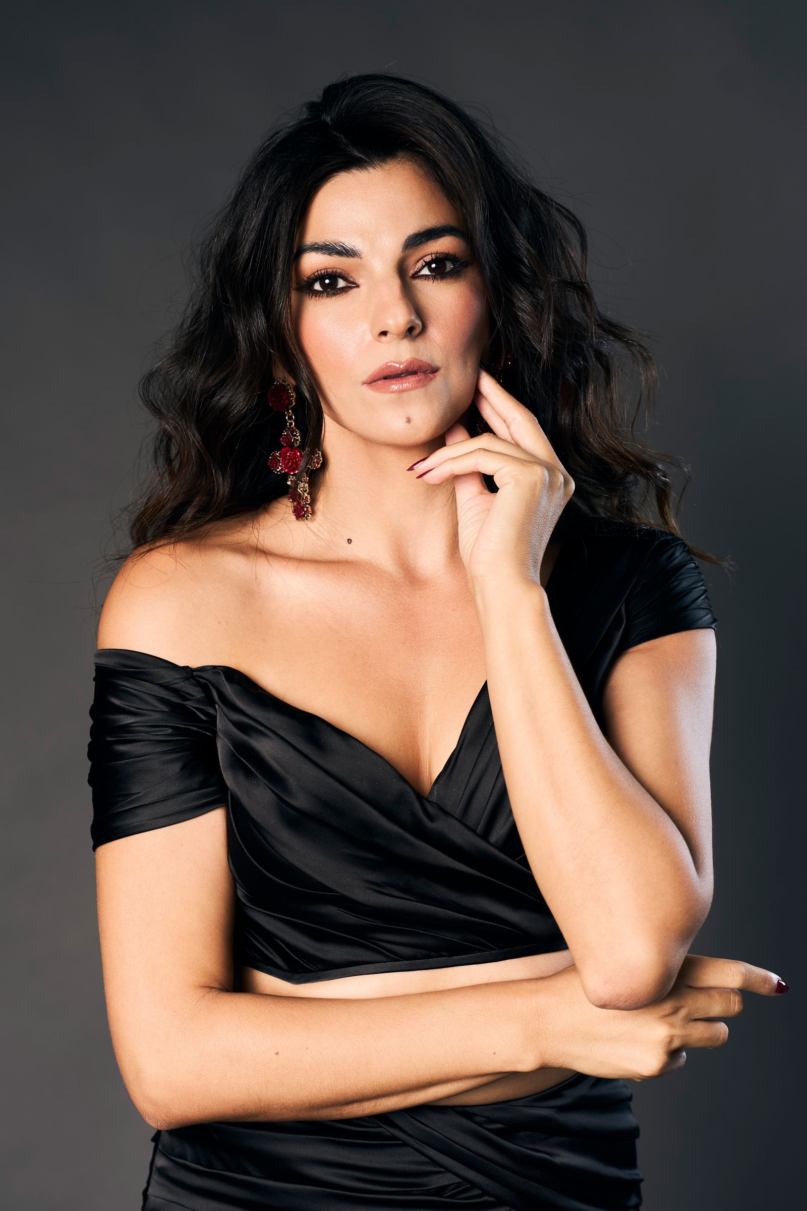 Dark wavy hair and a black off-the-shoulder bodice against a deep studio ground, hand raised to the jaw.
