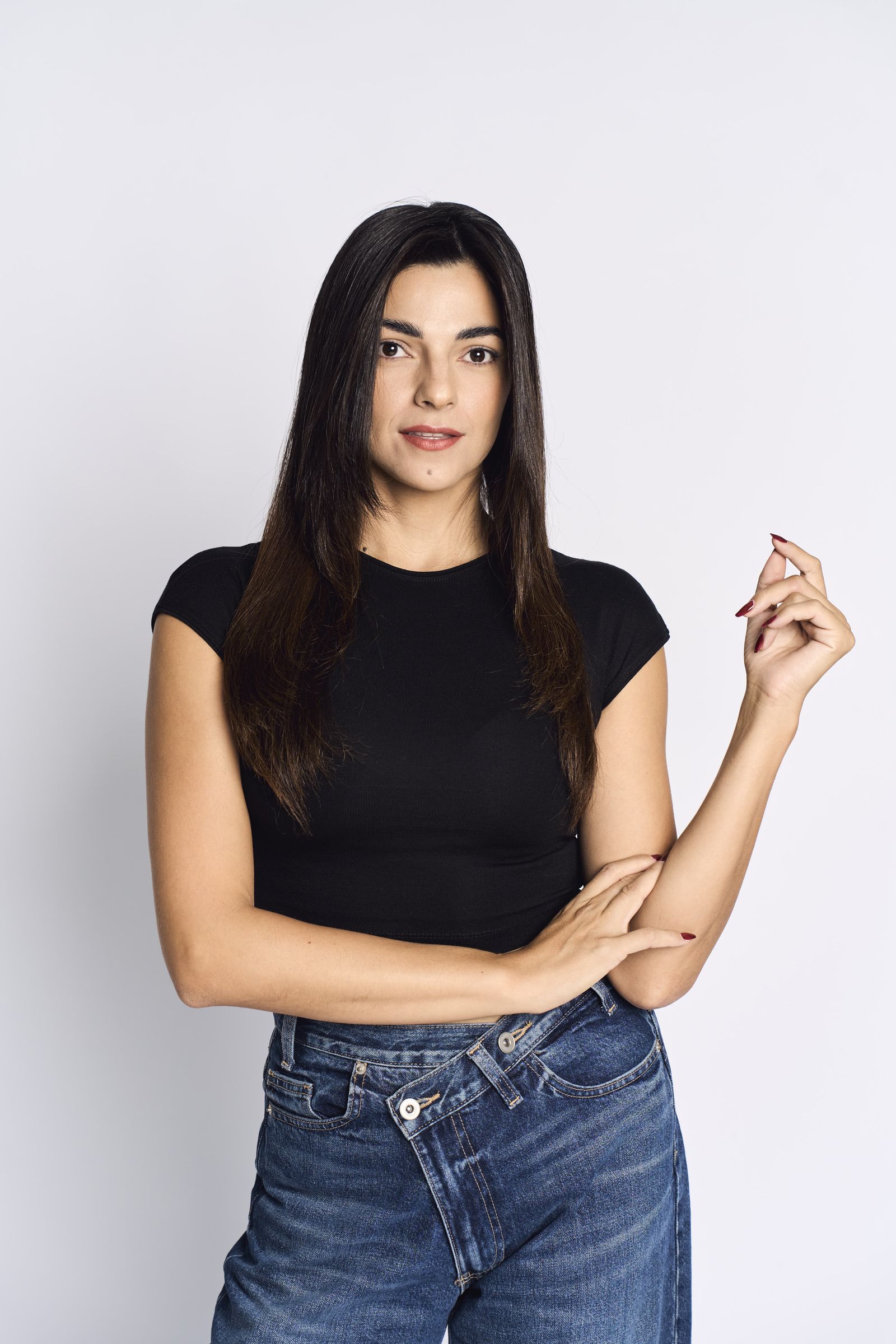 Black t-shirt and jeans, straight dark hair, arms crossed against a bright white background.