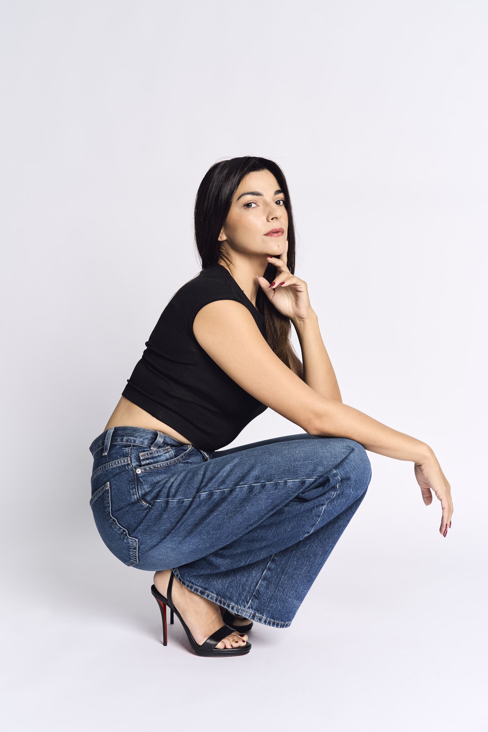 Wide-leg jeans and heels, crouched in profile against a white background under even light.