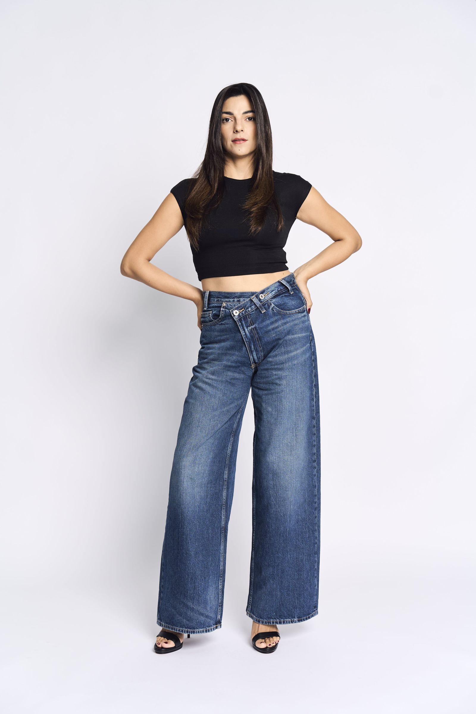 Standing full length in a black crop top and wide-leg jeans, hands on hips, white background.