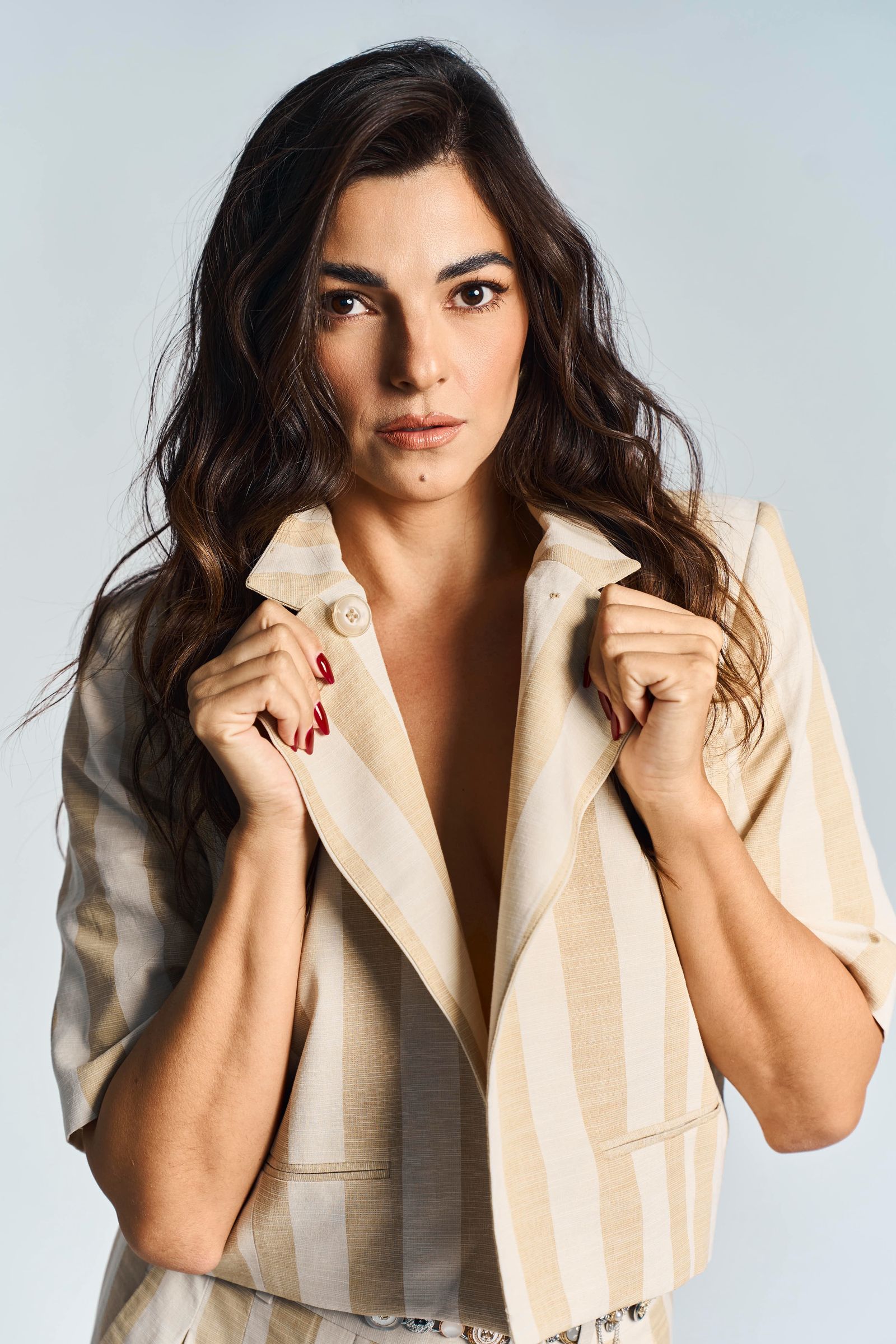 Close portrait, red nails holding the lapels of a striped jacket, looking straight down the lens.