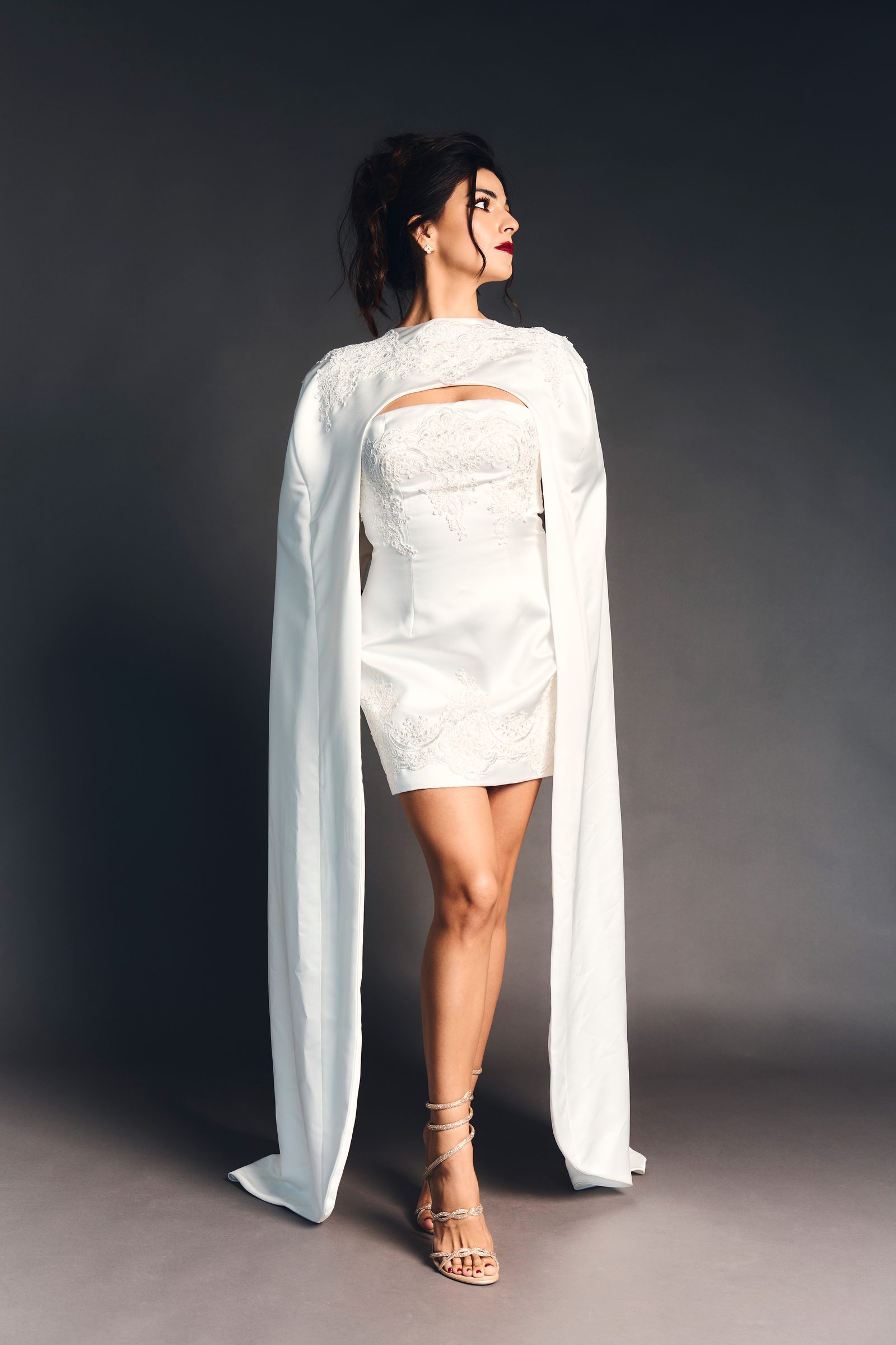 Full length in a white lace mini dress with a floor-length cape, turned away, looking back over the shoulder against a graphite ground.