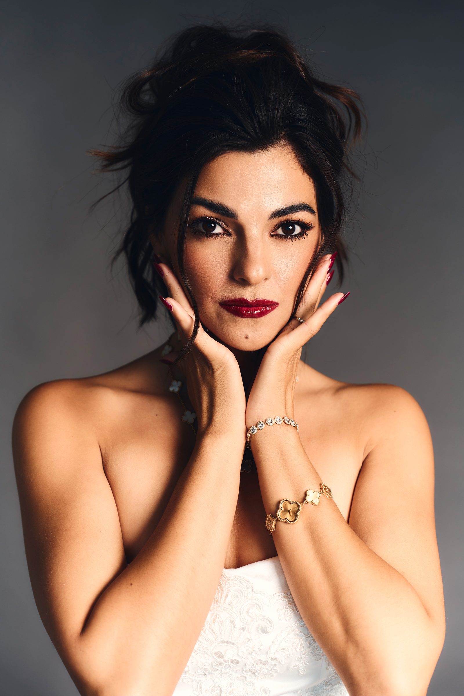 Beauty portrait with both hands framing the face, a gold bracelet and rings catching the light, red lip and swept-back hair.