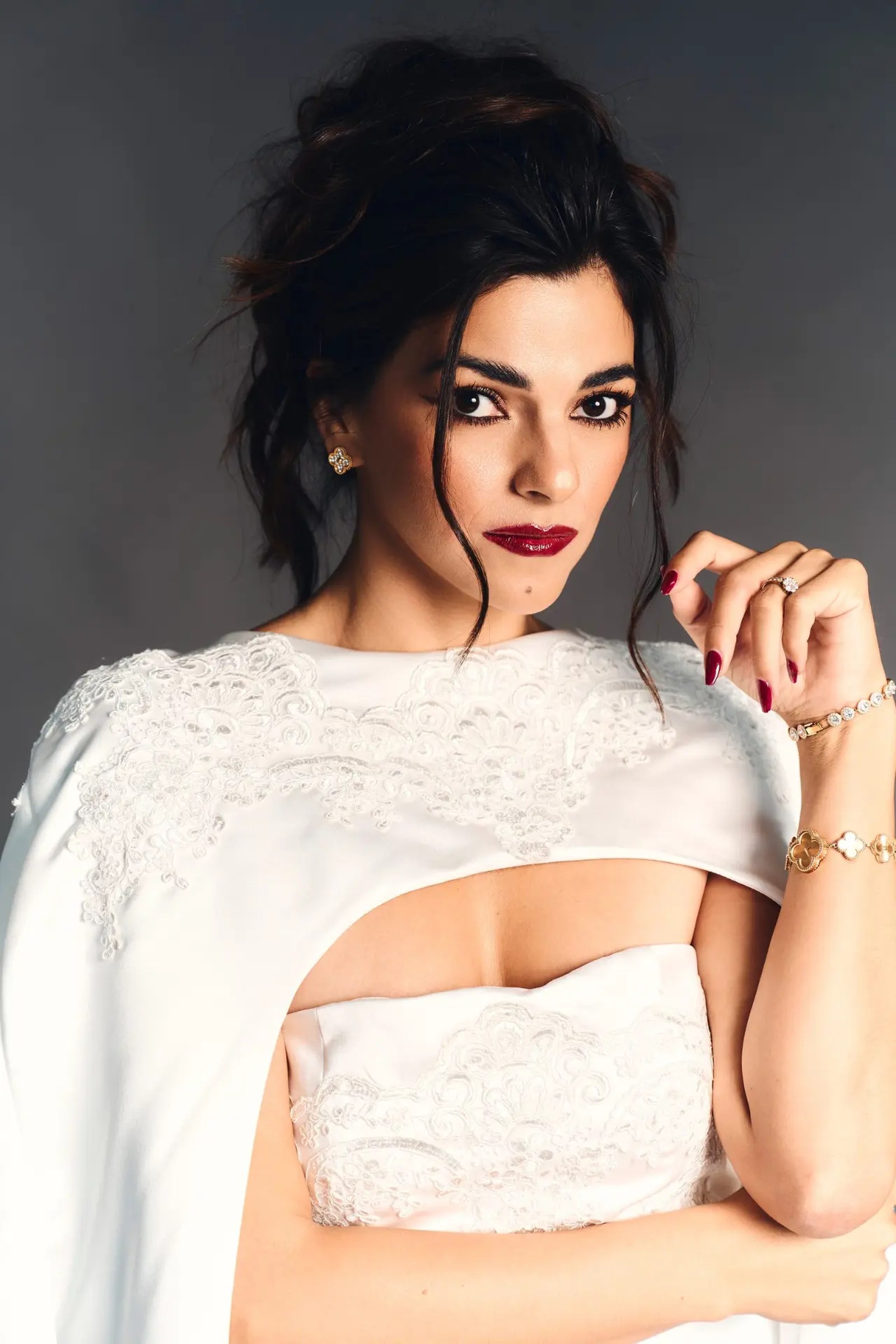 Portrait in an ivory lace-trimmed cape and bustier, hair swept up, gold and diamond jewellery, deep grey studio ground.