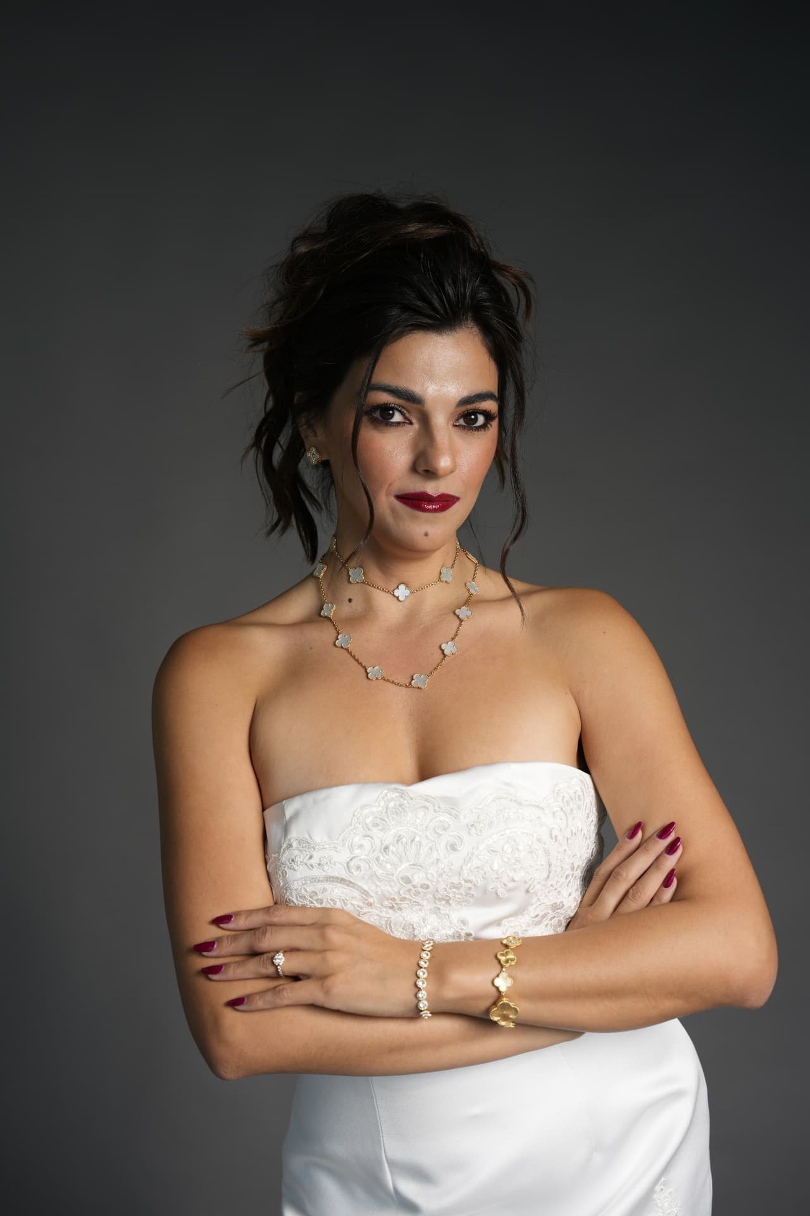Portrait in a strapless white satin and lace gown, arms folded, hair swept up, gold clover necklace and bracelets against a graphite ground.